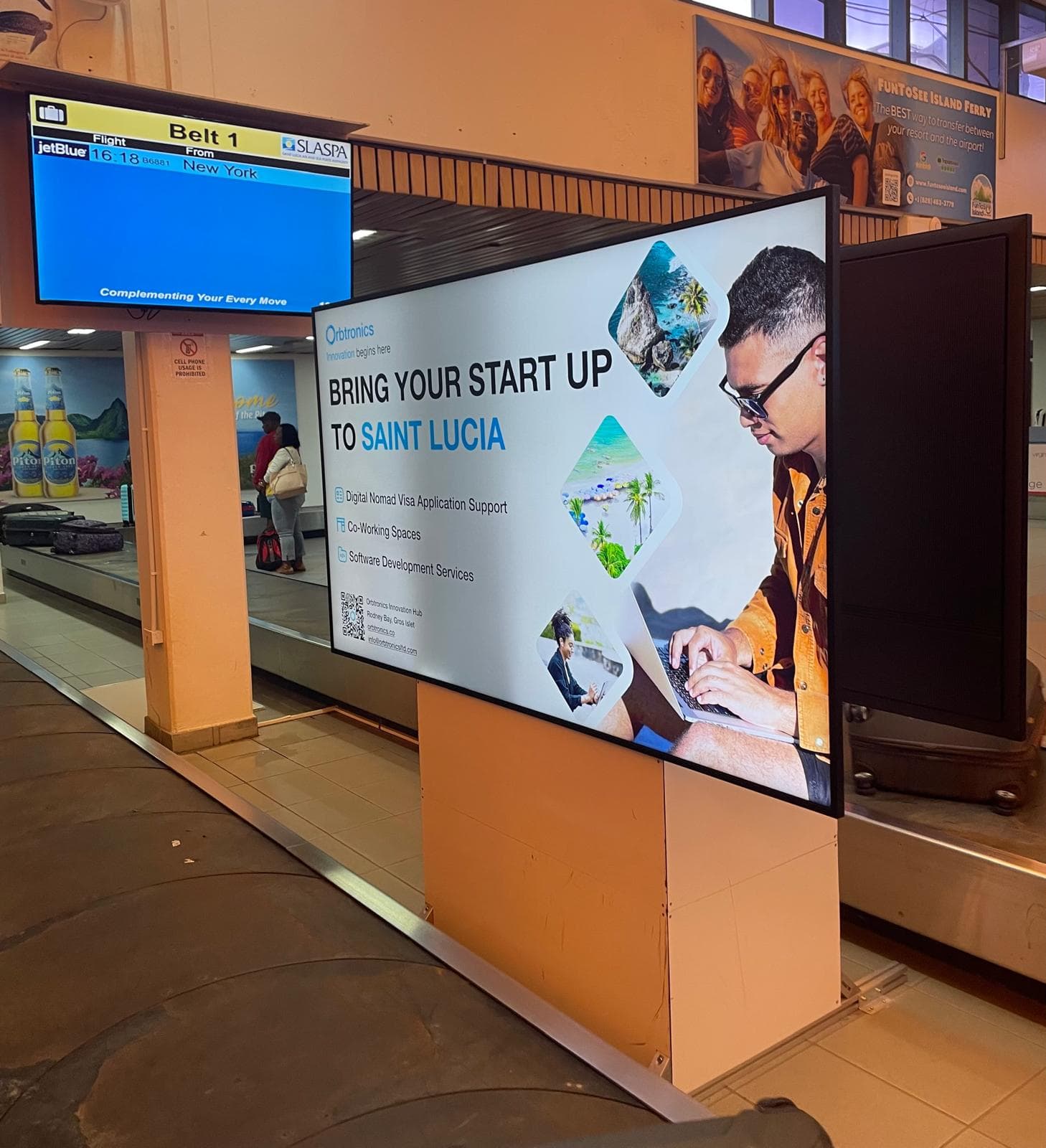 A digital advertisement for Orbtronics displayed at the baggage claim area in Hewanorra International Airport, Saint Lucia. The ad reads “Bring Your Start Up to Saint Lucia” and highlights services such as Digital Nomad Visa Application Support, Co-Working Spaces, and Software Development Services. The screen shows a man using a laptop, alongside scenic images of Saint Lucia.